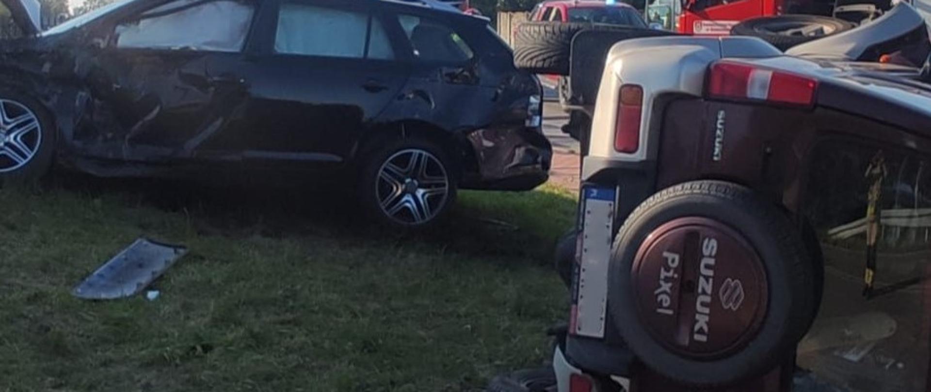 Zdjęcie przedstawia dwa uszkodzone samochody. Z lewej stoi na kołach Volkswagen, który uderzył w ogrodzenie, z prawej leży na boku Suzuki Jimmy. W głębi widać fragmenty samochodów pożarniczych.