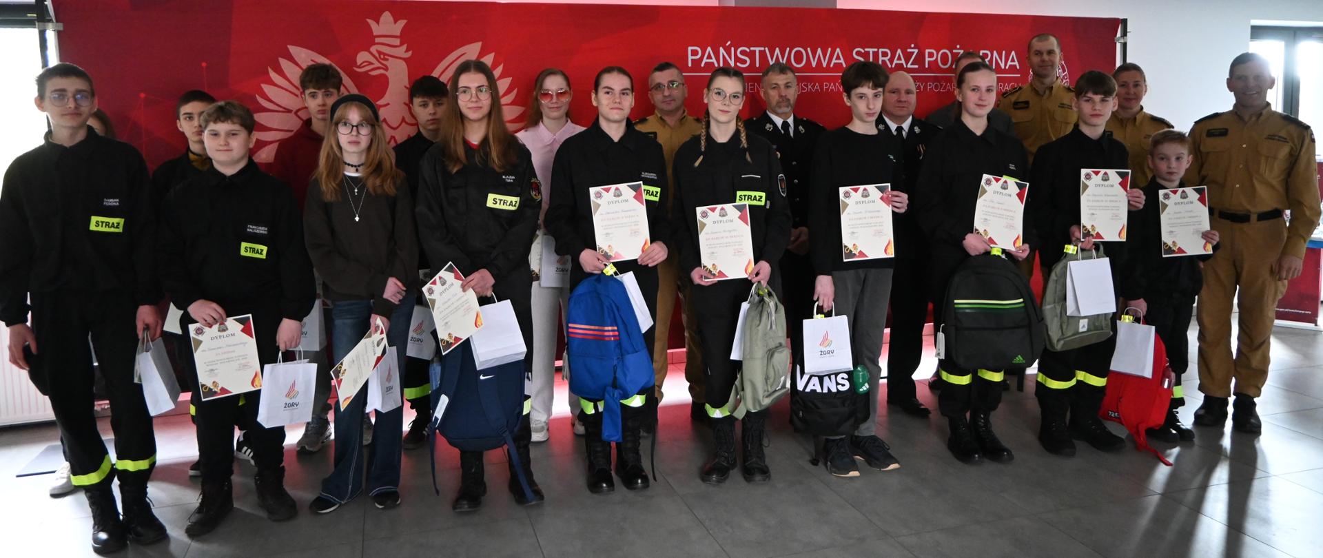 Grupa kilkunastu młodych osób stoi w rzędzie w pomieszczeniu, pozując do zdjęcia z dyplomami i torbami z nagrodami. Większość ma na sobie czarne stroje z naszywkami „STRAŻ”, a niektórzy elementy odblaskowe. Za nimi stoją dorośli w mundurach strażackich. W tle widoczny jest czerwony baner z napisem „Państwowa Straż Pożarna” oraz symbolem orła