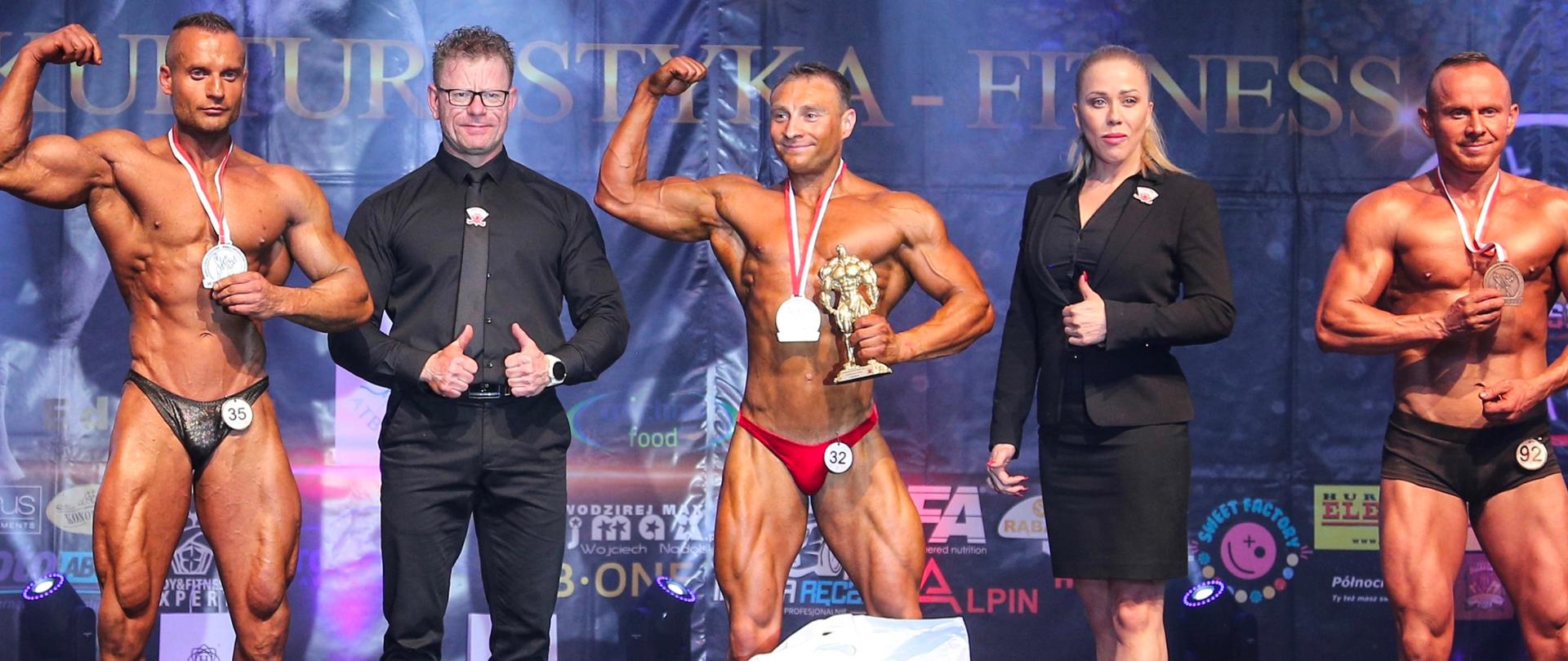 Na scenie, po wręczeniu nagród widoczni trzej zawodnicy kulturystyki oraz mężczyzna i kobieta, którzy stoją wśród nich. 