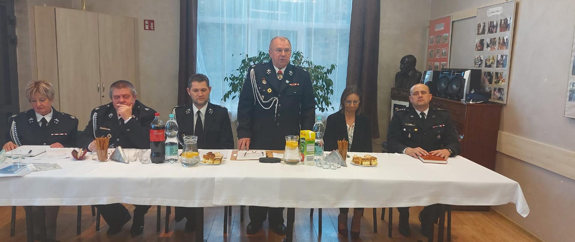 Na zdjęciu widać siedzących przy stole druhów i druhny OSP z Lutowisk w mundurach galowych. Z prawej strony widać również komendanta powiatowego PSP w Ustrzykach Dolnych. Na stole jest biały obrus przekąski i napoje. Z tyłu duże okno, po lewej szafa a po prawej komoda na której stoi wieża z głośnikami.