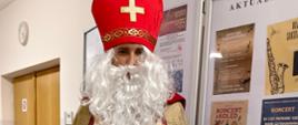 Osoba przebrana za Świętego Mikołaja (w biskupim stroju) stoi w szkolnym korytarzu, ubrana w białą albę, złoto-czerwony płaszcz, czerwoną mitrę i białą, kręconą brodę. Trzyma w dłoni dzwonek i czerwony woreczek, a obok widoczna jest gablota z informacjami.