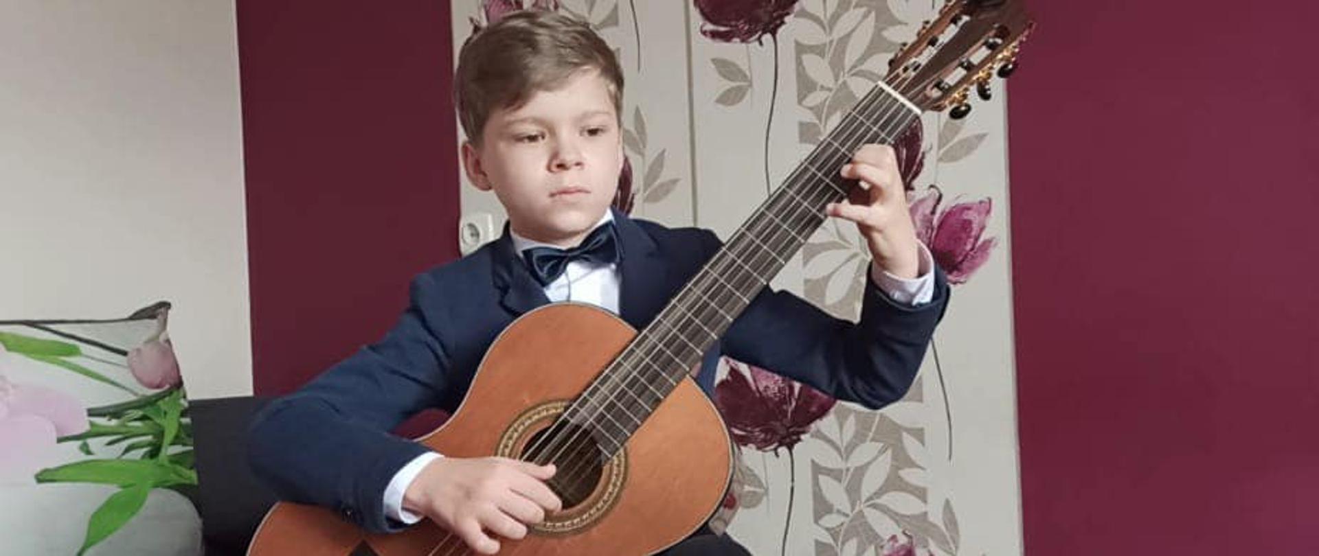 Chłopiec siedzi i trzyma gitarę, ubrany w stój galowy, za nim bordowa ściana z białym pasem pionowym, na nim kwiaty.