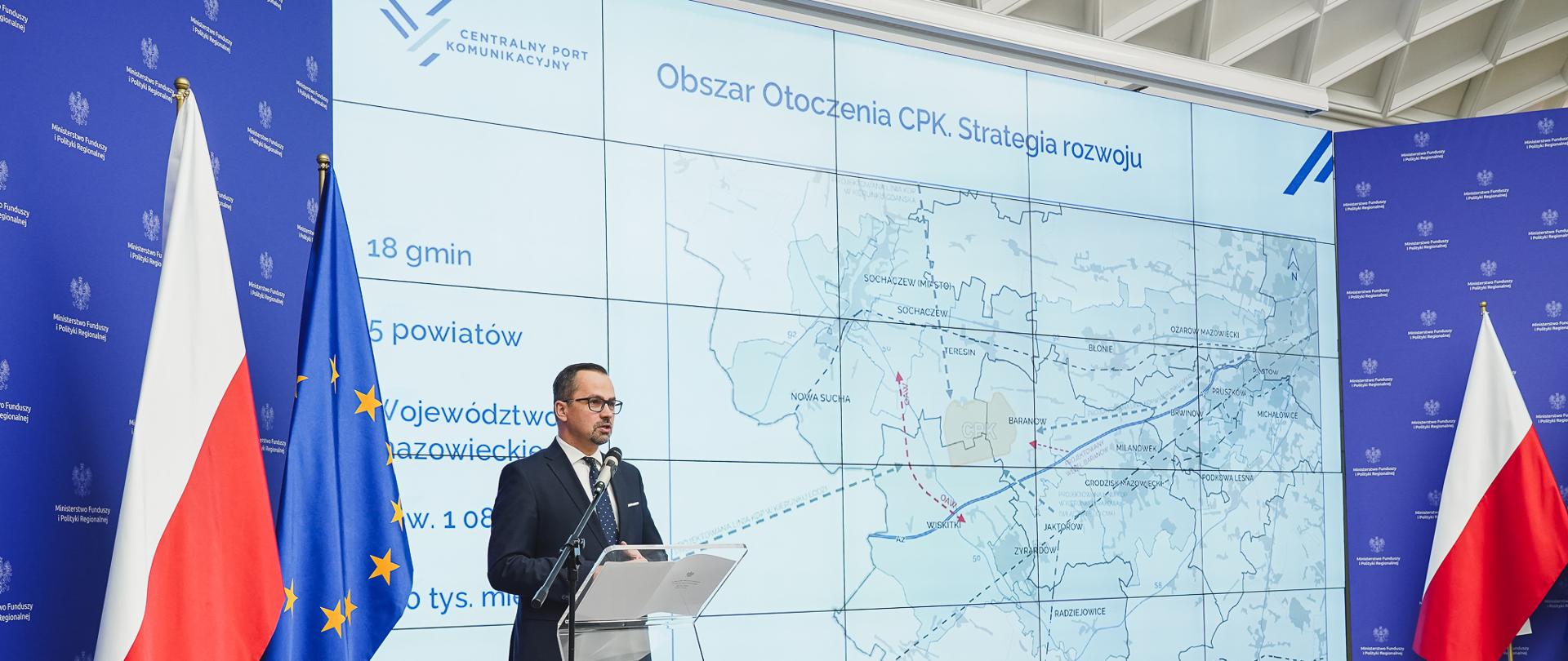 Minister Marcin Horała stoi na scenie przy mównicy i mówi do mikrofonu.. Po lewej stronie od niego stoi na statywie mikrofon oraz flagi Polski i Unii Europejskiej.