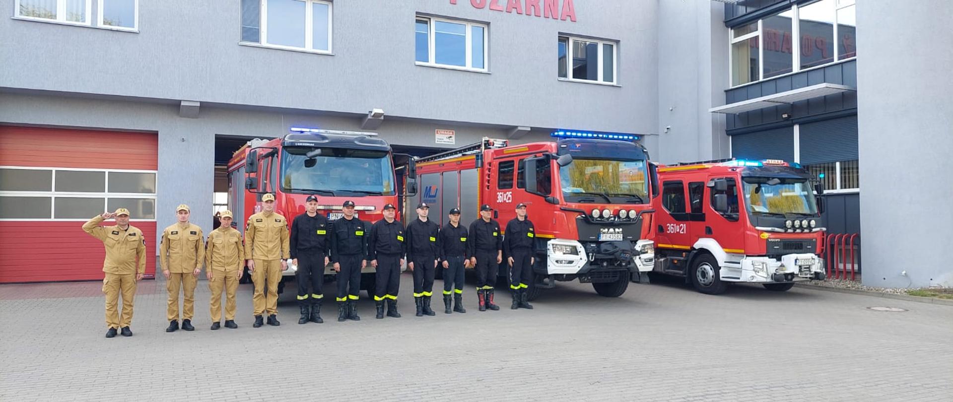  Strażacy stoją na placu przed remizą i minutą ciszy oddali hołd zmarłemu druhowi. Z tyłu widać samochód strażacki na włączonych sygnałach świetlnych i dźwiękowych
