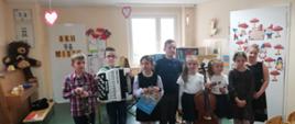 Zdjęcie przedstawia uczniów szkoły muzycznej stojących w jednym rzędzie przed przedszkolakami. Niektóre dzieci trzymają w rękach instrumenty muzyczne: akordeon, klarnet oraz wiolonczelę. 