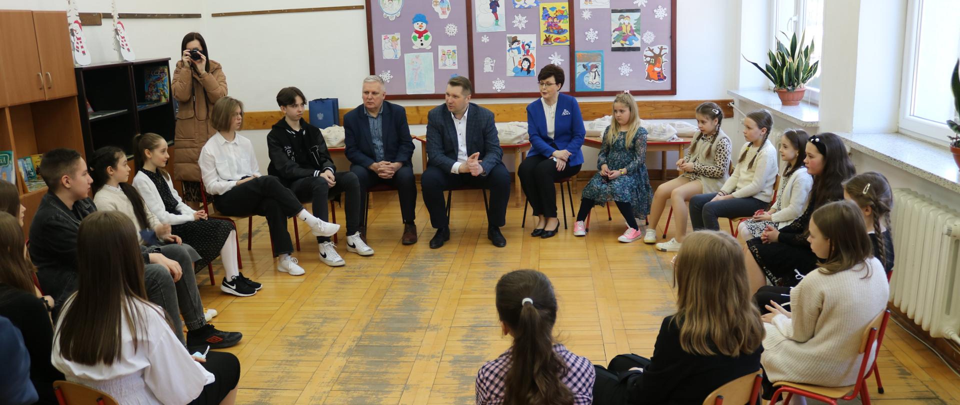 Minister siedzi na krześle, wśród dzieci ze szkoły. Krzesła ustawione są w koło.
