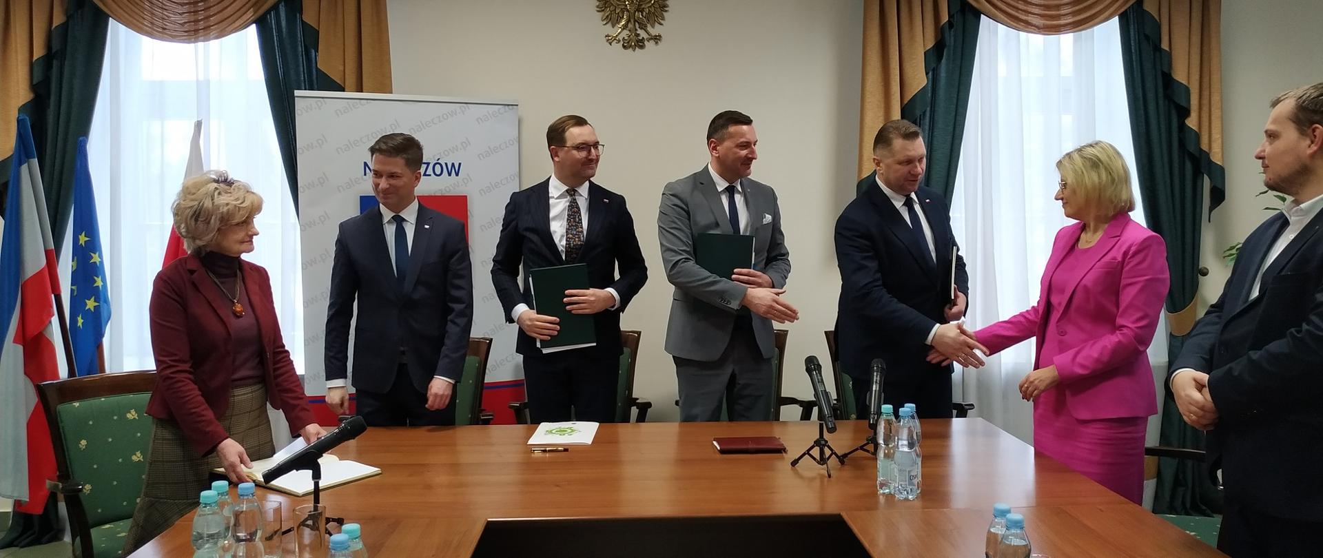 Przed ustawionymi w czworokąt drewnianymi stołami stoi pięciu mężczyzn w garniturach i dwie kobiety, jedna ubrana na czerwono stoi z boku, druga ubrana na fioletowo ściska rękę jednemu z mężczyzn, za nimi żółta ściana i dwa przysłonięte zasłonami okna.