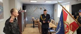 Zbiórka strażaków w jasnym pomieszczeniu. Po prawej stronie widoczny biało - czerwony sztandar komendy. Po lewej funkcjonariusz w mundurze koszarowym składa ślubowanie. W środku komendant w mundurze wyjściowym odczytuje rotę ślubowania z granatowej teczki. W tle wyposażenie gabinetu.