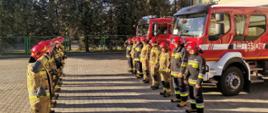 Zdjęcie przedstawia dwie zmiany służbowe - osiemnastu strażaków stojących przed budynkiem komendy straży w wieluniu podczas minuty ciszy dla ukraińskich strażaków, za nimi stoją dwa samochody bojowe