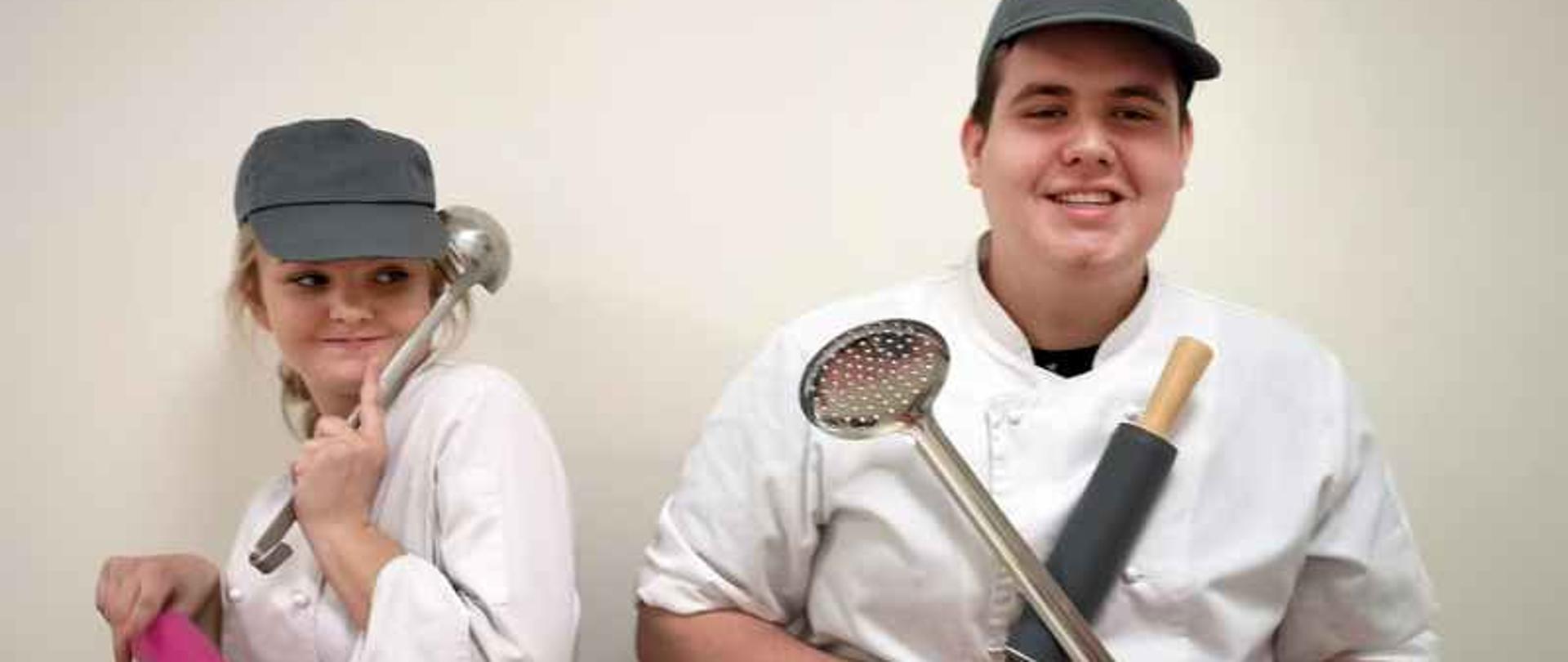 Na zdjęciu jest chłopak i dziewczyna w strojach kucharzy. Dziewczyna trzyma wazówkę i wałek a chłopak ma w rękach wałek i cedzak.