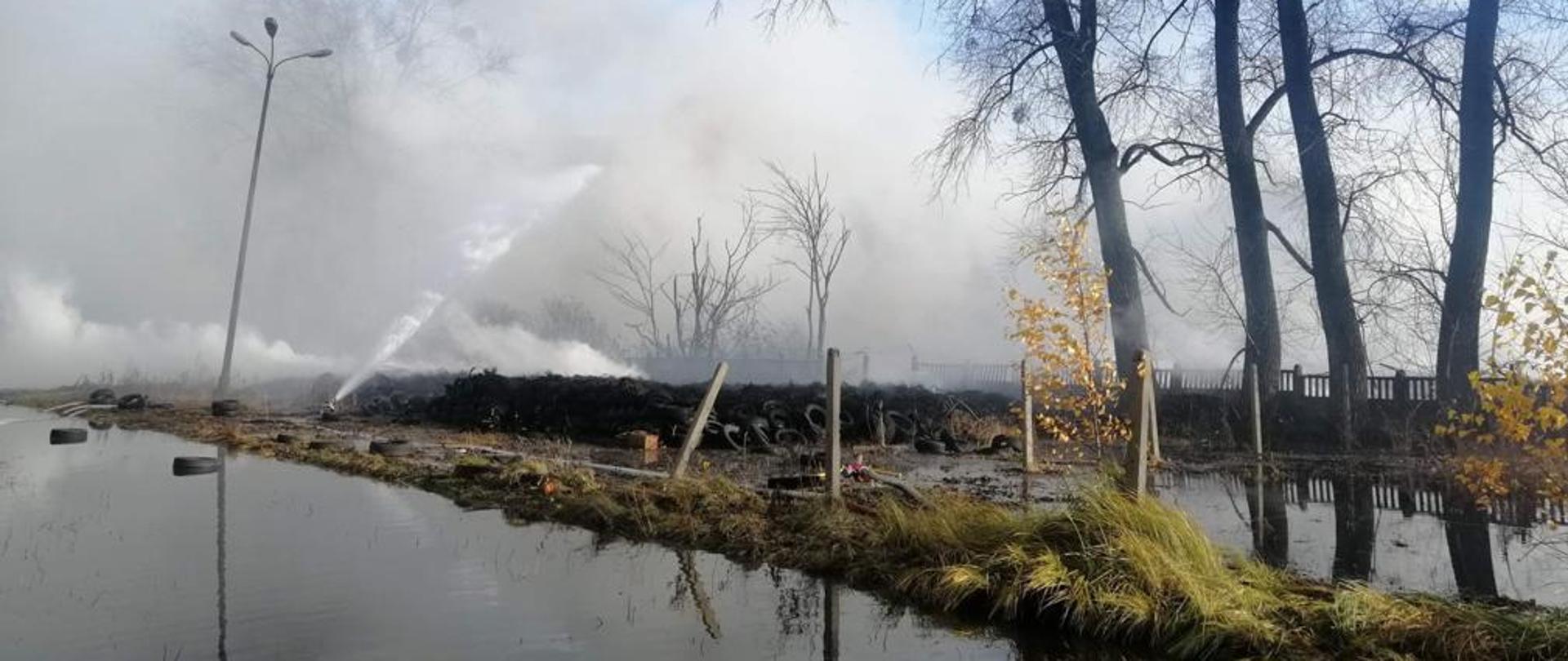 Zdjęcie częściowo ugaszonego pożaru składowiska opon w Raciniewie, widoczne linie gaśnicze oraz podawane prądy wody, w tle unoszący się biały i szary dym.