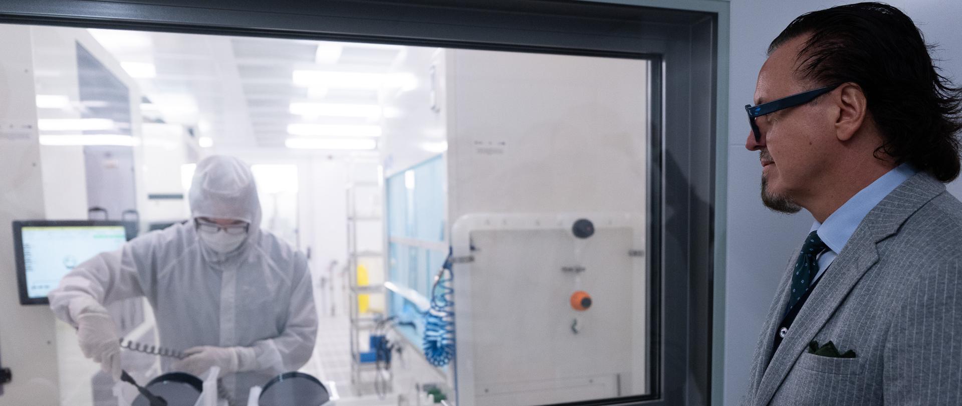 Minister Balczun z wizytą w Centrum Zaawansowanych Materiałów i Technologii CEZAMAT Politechniki Warszawskiej. Minister stoi przed szybą, za którą widać osobę ubraną w kombinezon roboczy podczas pracy. 