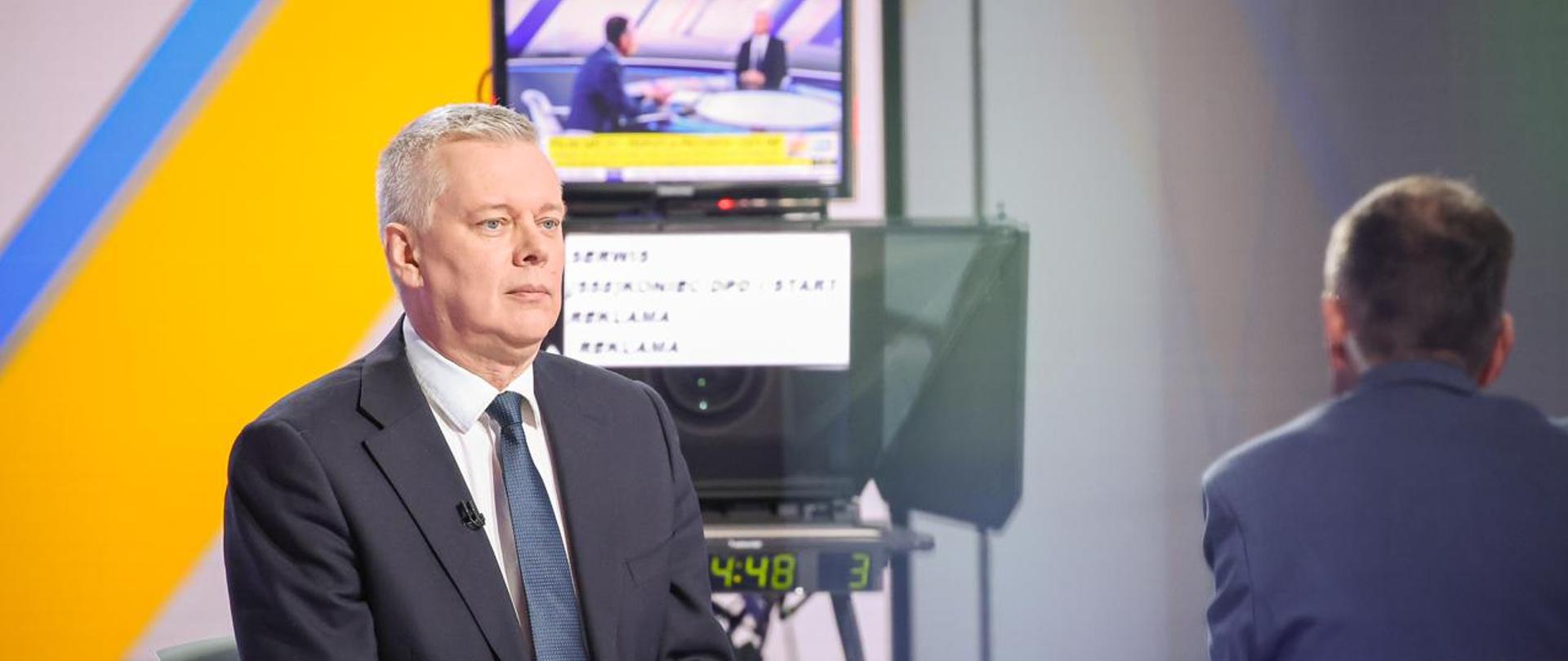 Po lewej stronie minister Tomasz Siemoniak, po prawej redaktor Konrad Piasecki. Minister zasiada przy stole, na którym znajduje się szklanka z wodą. W tle, w centralnym punkcie, kamera z monitorem. Na monitorze wyświetla się zdjęcie ww. osób. Po lewej stronie w tle ekran z żółtym szerokim paskiem. 