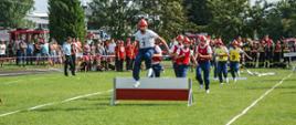 Na zdjęciu strażacy w strojach sportowych biegnący po torze do ćwiczenia bojowego, w tle kibice obserwujący bieg zza taśmy ostrzegawczej.