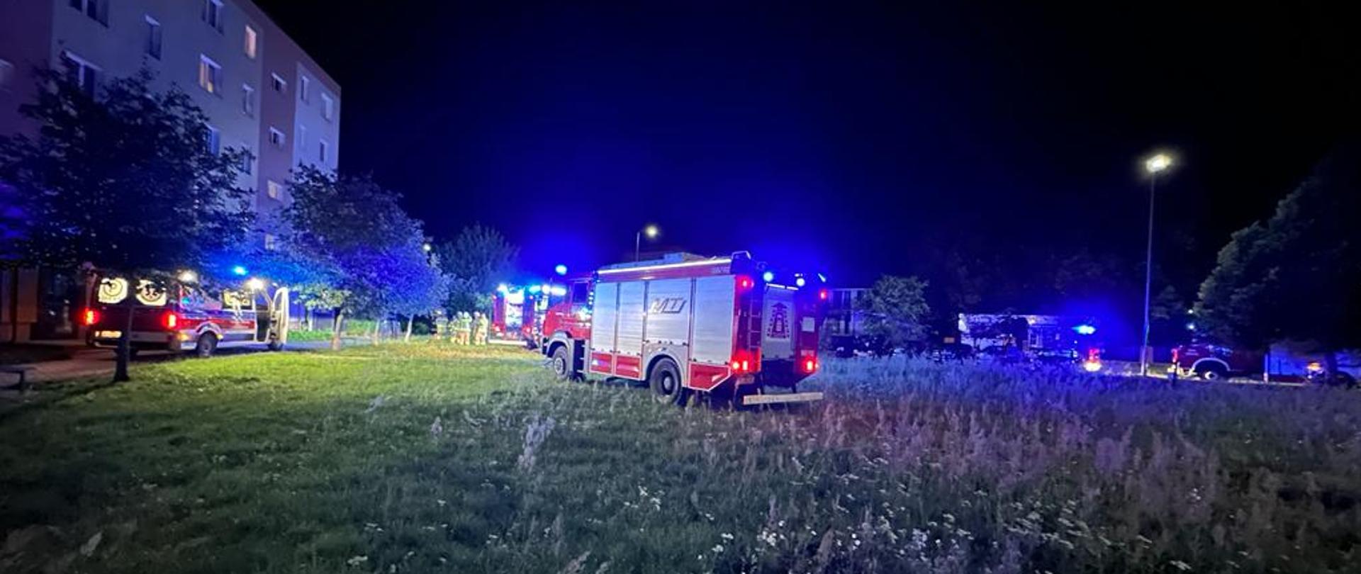 Zdjęcie przedstawia dwa pojazdy pożarnicze oraz ambulans podczas zdarzenia związanego z ugaszeniem pożaru mieszkania na I piętrze budynku wielorodzinnego