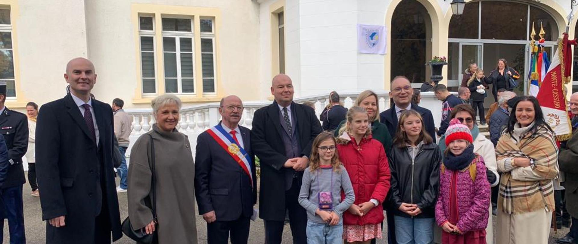 Na zdjęciu dorosłe osoby elegancko ubrane i czworo dzieci stoją przed budynkiem hotelu, obok osoby trzymające sztandary.