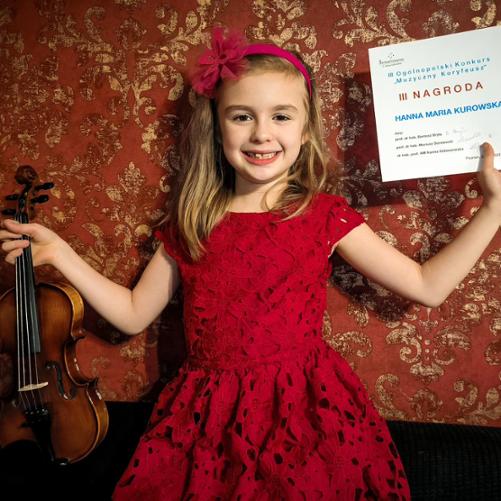 Postać dziewczynki, która w jednej ręce trzyma skrzypce, a w drugiej podniesionej do góry trzyma swój dyplom.