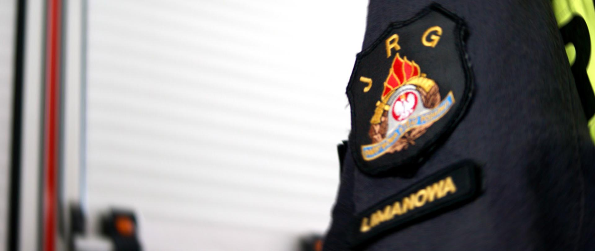 Naszywka - logo JRG Limanowa na ubraniu specjalnym w garażu na tle wozu strażackiego PSP Limanowa.