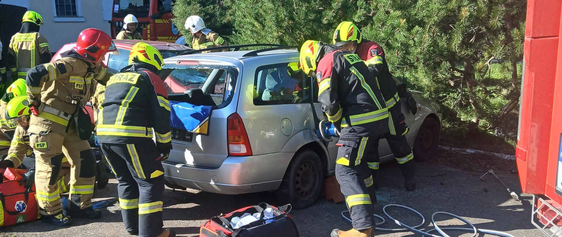 Strażacy w hełmach i ubraniach bojowych prowadzą działania ratownicze przy rozbitym samochodzie osobowym