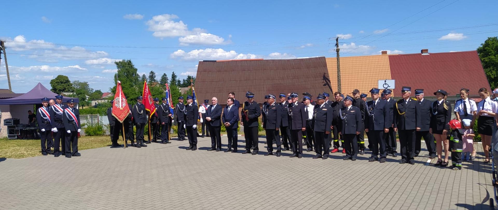 Strażacy i goście czekają na rozpoczęcie uroczystości na placu przed remiza Ochotniczej Straży Pożarnej w Podróżnej. W tle strażacy w umundurowaniu ustawieni w dwuszeregu, sztandary sąsiednich jednostek straży oraz zaproszeni goście. 