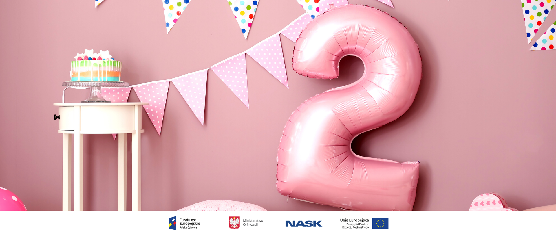 Duży różowy balon w kształcie cyfry 2. Obok stolik, na którym stoi kolorowy tort.