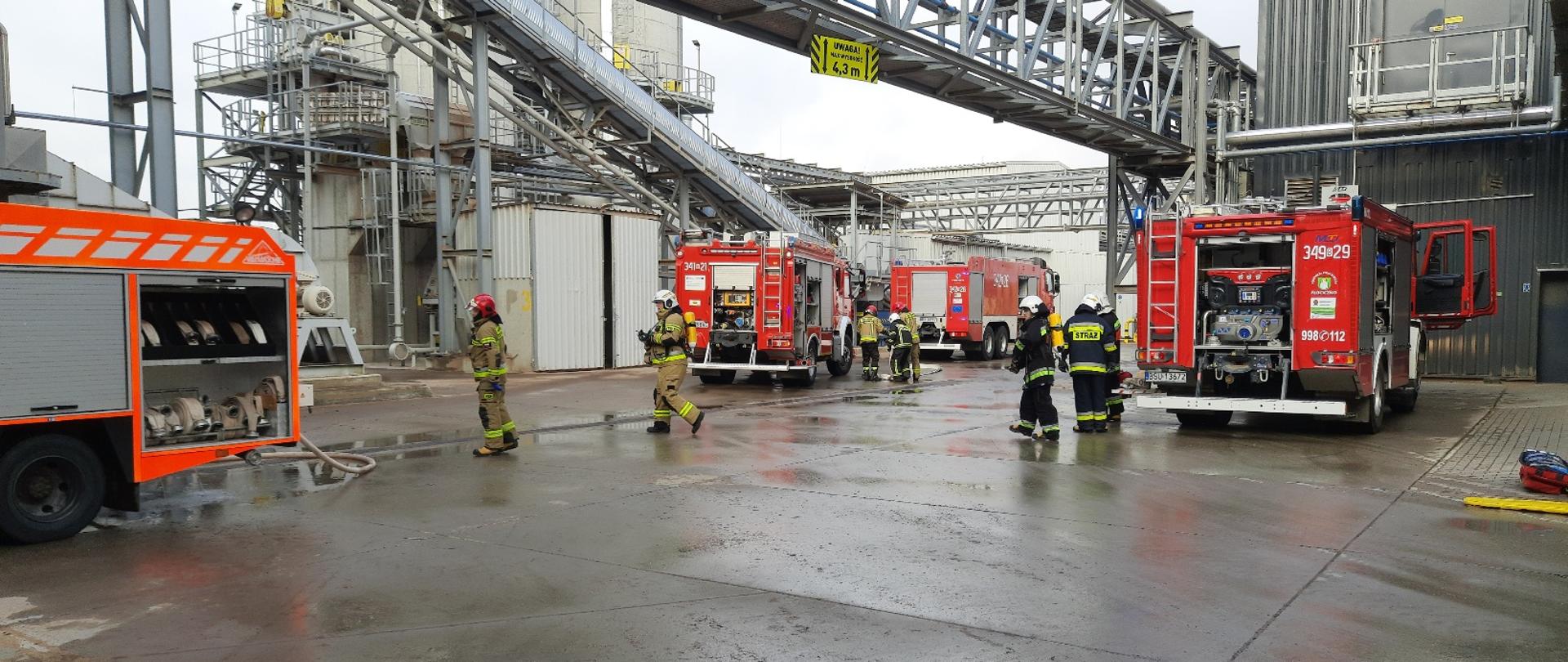 Na zdjęciu widać strażaków i samochody pożarnicze w tle budynki fabryki