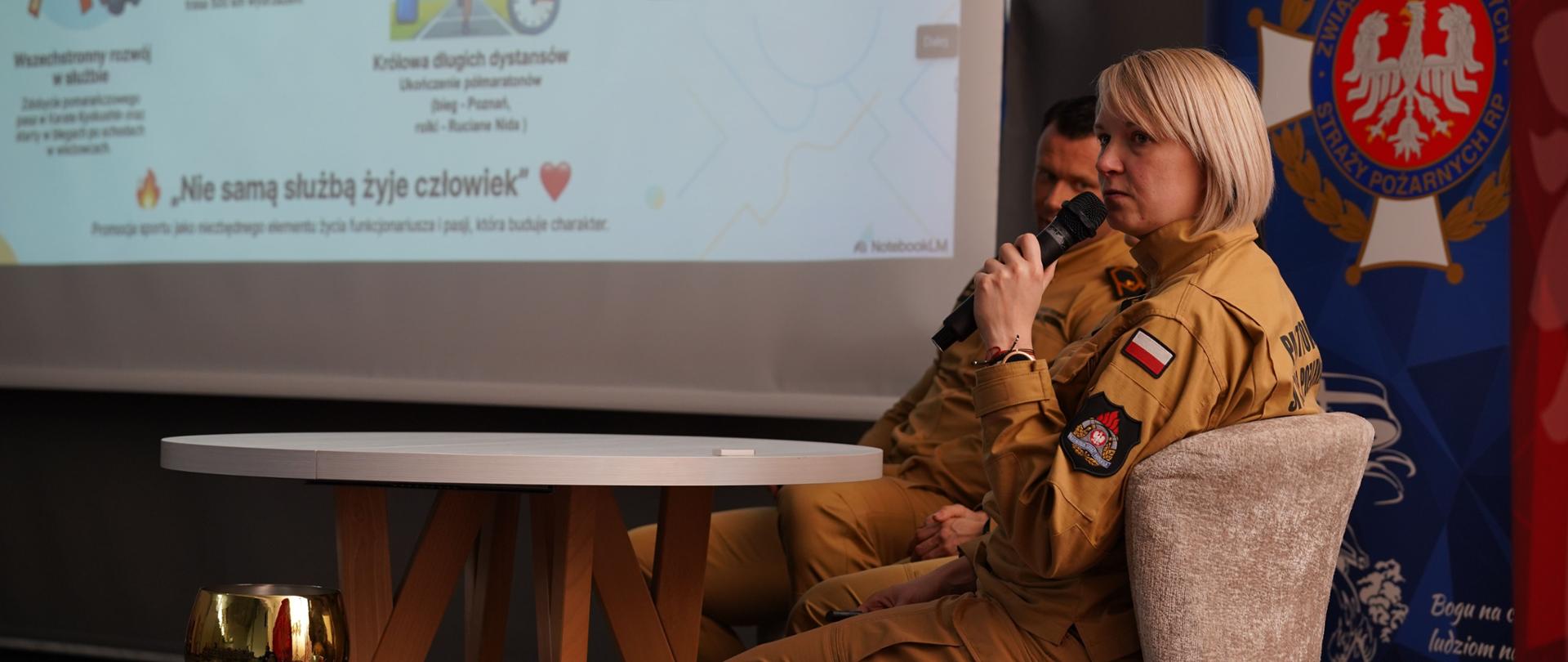 Strażaczka w mundurze siedzi na krześle i trzyma mikrofon, przemawiając podczas spotkania lub prezentacji. Obok niej, częściowo widoczny, siedzi inny strażak. W tle znajduje się ekran z prezentacją zawierającą ilustracje i teksty dotyczące aktywności fizycznej oraz służby, a także baner Ochotniczej Straży Pożarnej. Na pierwszym planie stoi stolik, a obok niego widoczny jest puchar i inne nagrody.