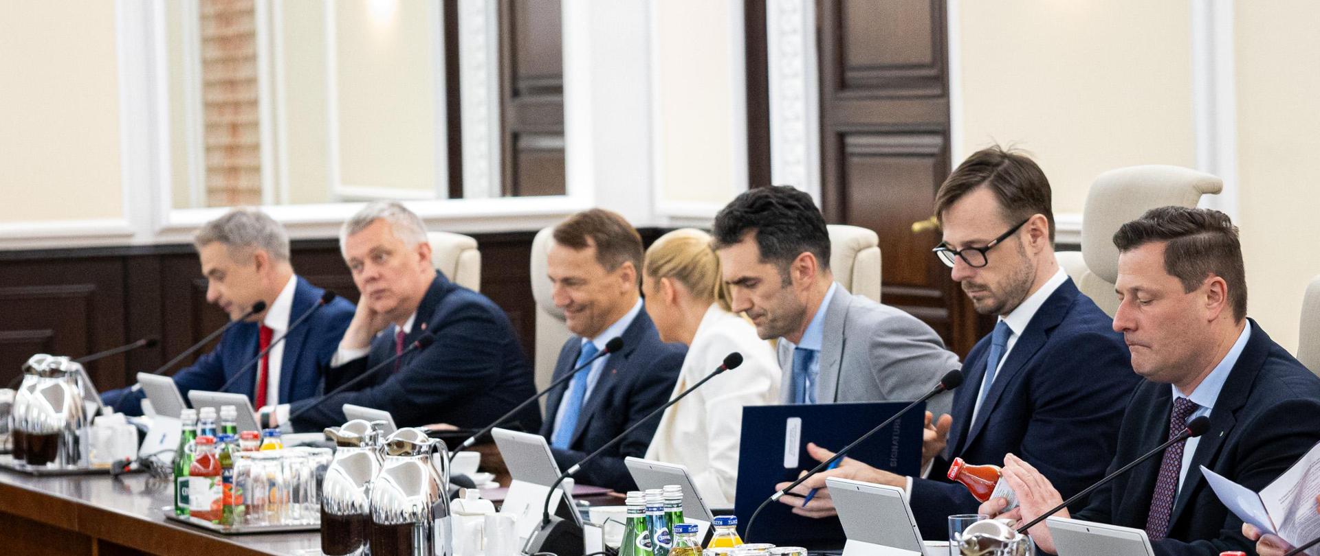 Rada Ministrów. Szeroki kadr. Przy drewnianym stole do obrad siedzi sześciu mężczyzn w garniturach i jedna kobieta w białej marynarce. Na stole laptopy oraz dzbanki do kawy i napoje w butelkach.