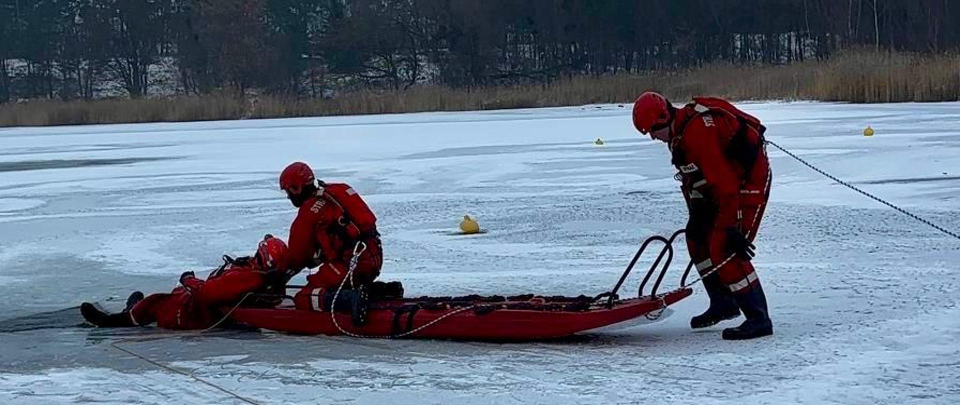 Dwóch ratowników ciągnie sanie ratownicze po lodzie, na których leży uratowana osoba, podczas gdy trzeci ratownik zabezpiecza akcję liną.