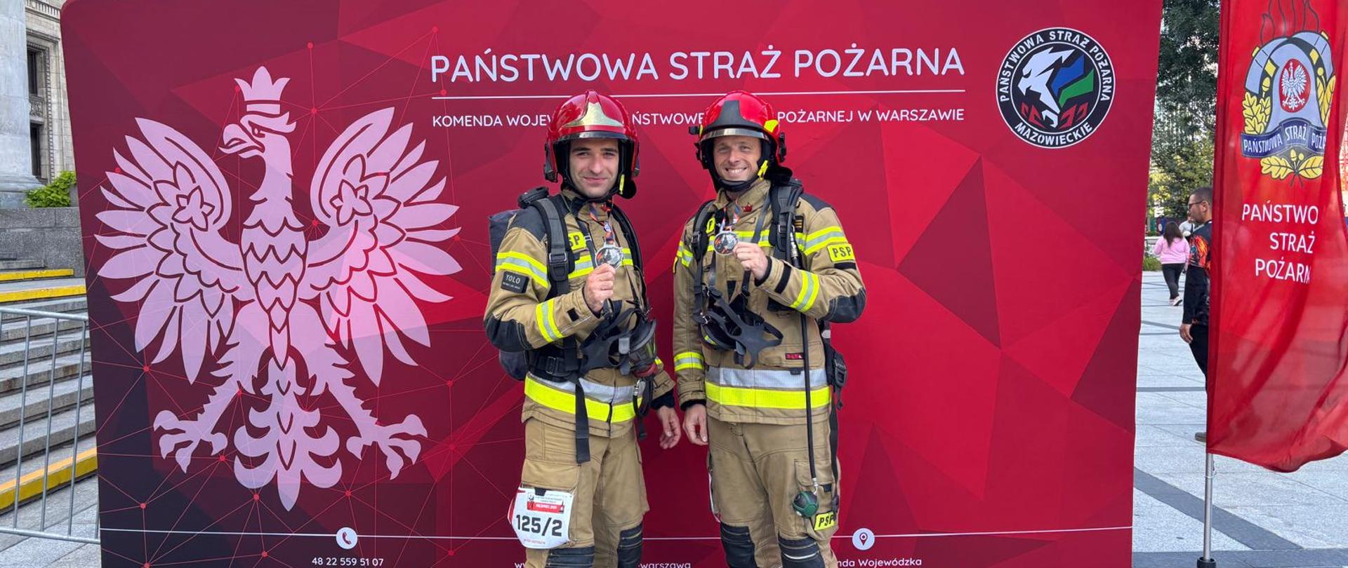 Strażacy na tle baneru Państwowej Straży Pożarnej. Jest to sportowa rywalizacja strażaków w biegu po schodach. Strażacy w umundurowaniu bojowym pozują po skończonym biegu