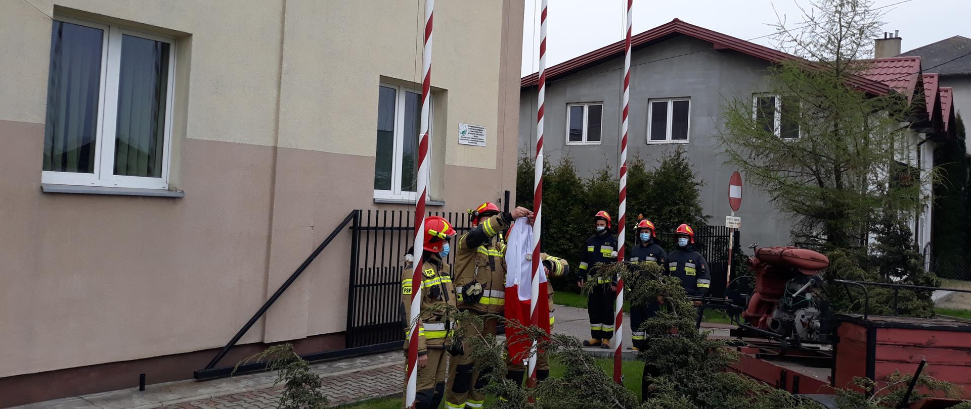 Przed budynkiem komendy szóstka strażaków ubranych w ubrania specjalne stoją, aby uroczyście zawiesić flagę państwową przed komendą. Jeden ze strażaków zawiesza flagę na maszcie.