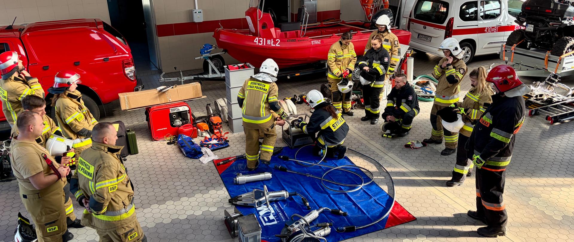 Zajęcia w hali garażowej strażnicy z zakresu ratownictwa technicznego. Kursanci w pełnym umundurowaniu bojowym i kaskach zgromadzili się wokół rozłożonego na macie hydraulicznego zestawu ratowniczego. W tle widoczne są pojazdy i sprzęt ratowniczy.
