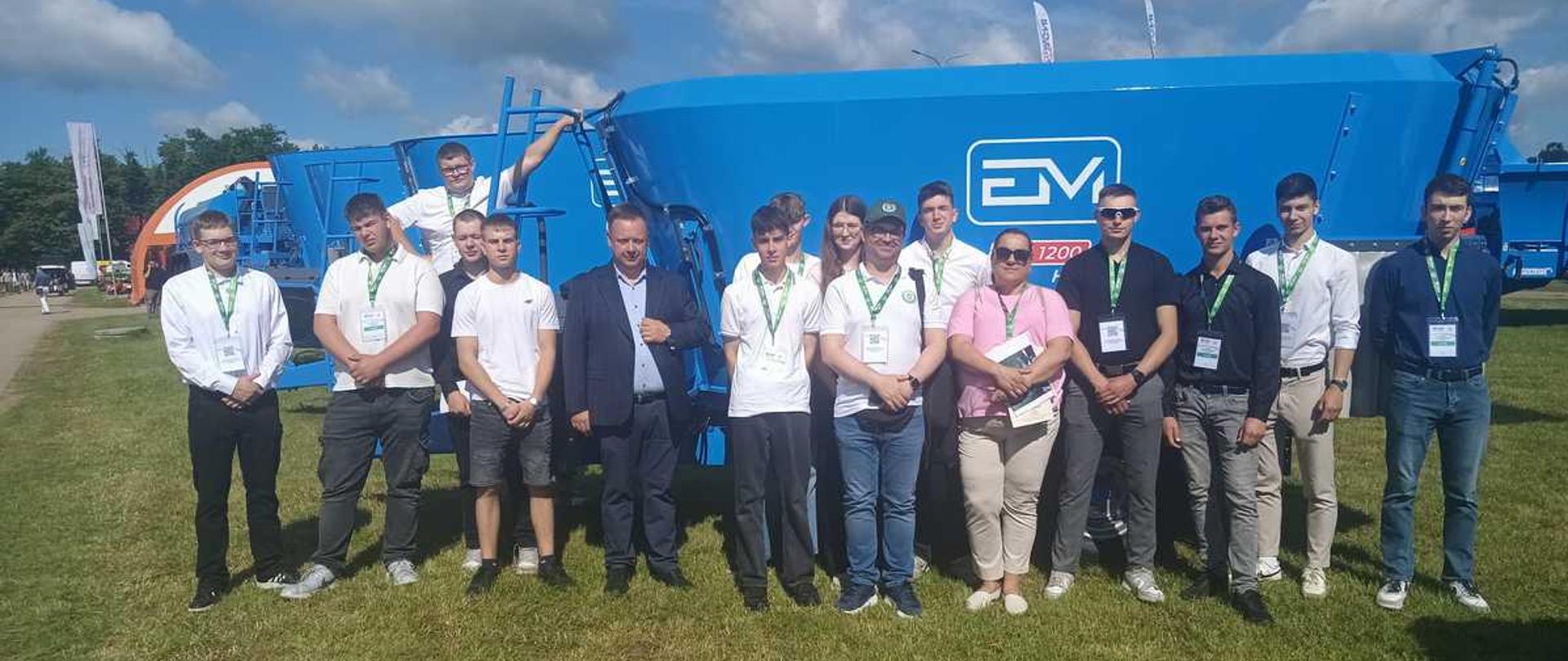 Zdjęcie przedstawia grupę czternastu osób pozujących na tle dużej, niebieskiej maszyny rolniczej pod bezchmurnym niebem.Opis ogólnyNa trawiastym placu, w pełnym słońcu, stoi rząd ludzi – głównie młodych mężczyzn oraz dwie kobiety. Wszyscy są ustawieni obok siebie i patrzą w stronę obiektywu. Za nimi wznosi się potężny, jasnoniebieski wóz paszowy marki EM.Osoby na zdjęciuSkład grupy: Większość stanowią nastolatkowie lub młodzi dorośli, prawdopodobnie uczniowie lub studenci. W środku stoi starszy od nich mężczyzna w ciemnej marynarce i białej koszuli.Ubiór: Dominują eleganckie, białe koszule z krótkim lub długim rękawem oraz ciemne spodnie (jeansy, chinosy). Kilka osób ma na sobie luźniejsze koszulki polo lub T-shirty.Identyfikatory: Prawie wszyscy uczestnicy mają na szyjach zielone smycze z zawieszonymi identyfikatorami, co sugeruje udział w zorganizowanym wydarzeniu, np. targach rolniczych.Tło i otoczenieMaszyna: Główny element tła to niebieski, metalowy zbiornik maszyny rolniczej. Po prawej stronie widoczne jest białe logo z literami „EM” oraz mniejszy napis „1200”. Z lewej strony konstrukcji zamontowana jest drabinka, na której opiera się jeden z chłopaków.Otoczenie: Grupa stoi na zielonej, lekko wysuszonej trawie. W oddali po lewej stronie majaczą inne maszyny, flagi reklamowe oraz ściana lasu. Na niebie widać pojedyncze, białe chmury. Ciepłe światło słoneczne rzuca wyraźne cienie na ziemię przed fotografowanymi osobami.