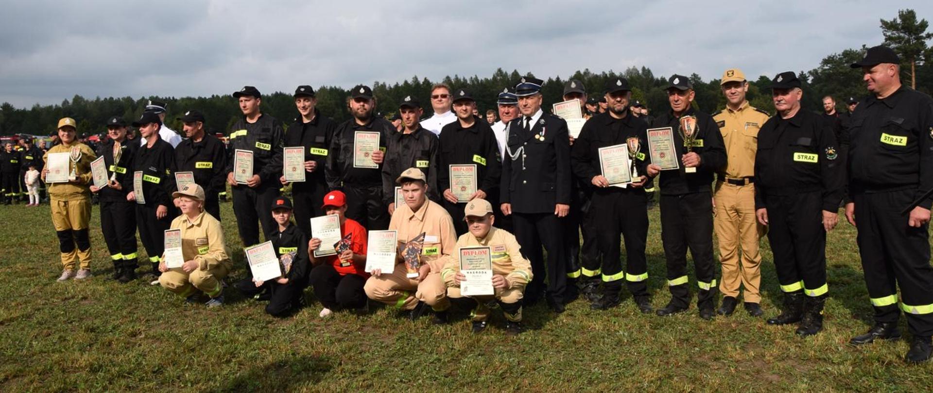 Zdjęcie przedstawia druhów z jednostek ochotniczych straży pożarnych i młodzieżowych drużyn pożarniczych w umundurowaniu koszarowym koloru czarnego i piaskowego. Zdjęcie wykonane na łące, w tle las iglasty. Pogoda pochmurna.