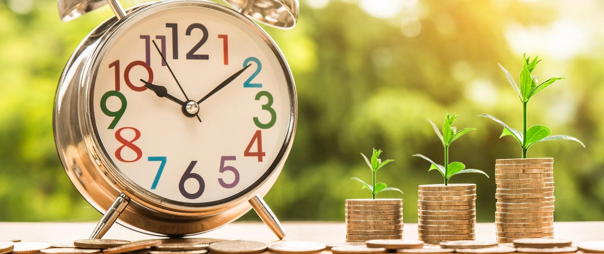 Na zdjęciu po lewej stronie okrągły zegarek, wskazujący na godzinę 10:10. Obok, na tle zieleni, trzy "kupki" monet, które ustawione są od najmniejszej do największej. Na każdej z nich widoczny jest wyrastająca z monet gałązka. 