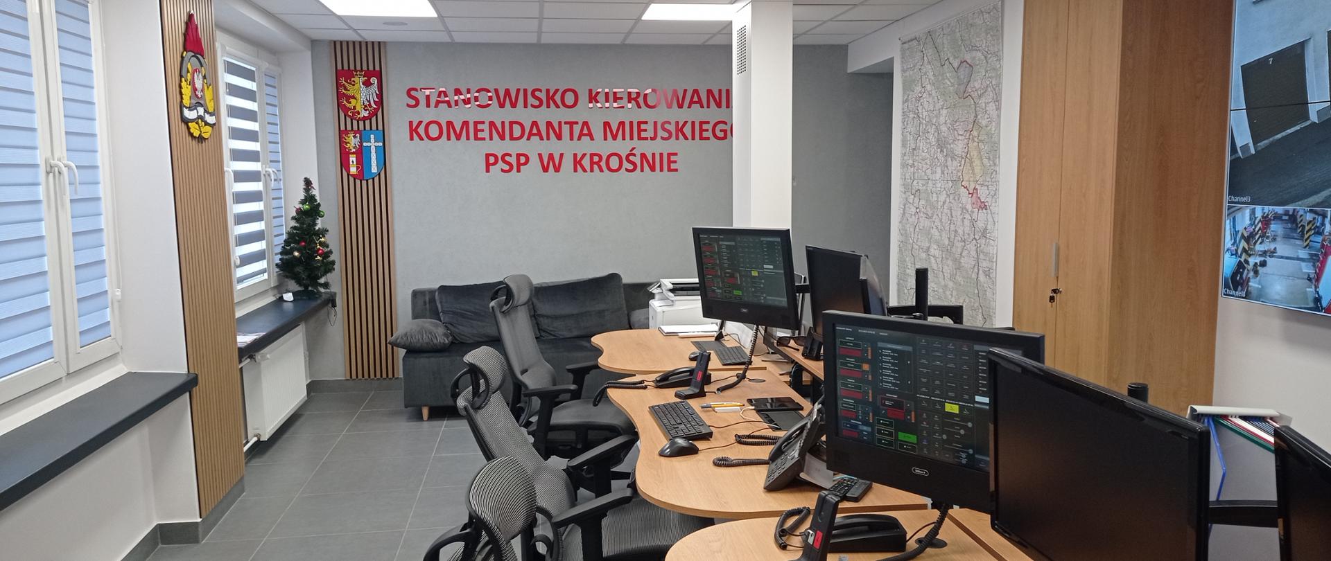 Zdjęcie przedstawia pomieszczenie stanowiska kierowania Komendanta Miejskiego PSP w Krośnie. W centrum zdjęcia znajdują się trzy stanowiska pracy dyspozytorów. Każde stanowisko wyposażone jest w fotel, dwa monitory oraz niezbędny sprzęt do alarmowania strażaków państwowej i ochotniczej straży pożarnej. 