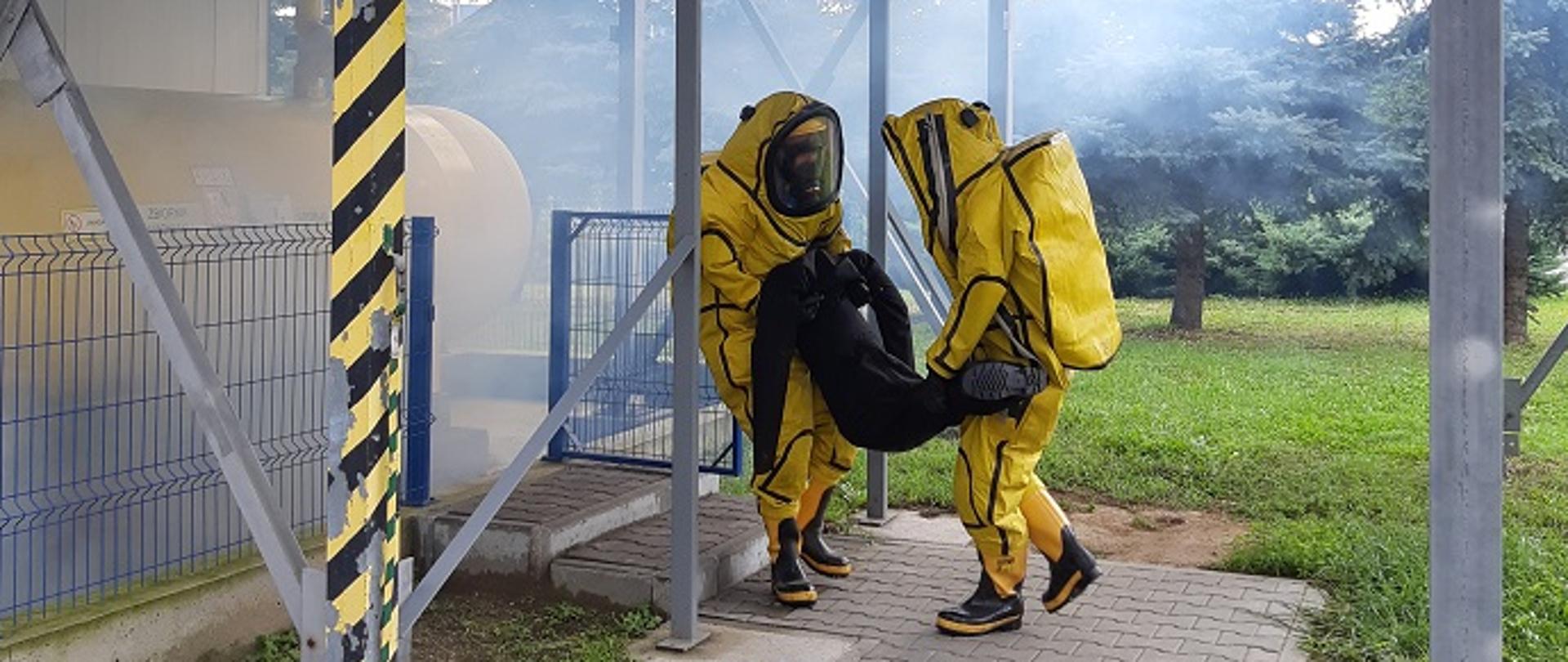 Na zdjęciu widzimy strażaków w ubraniach gazoszczelnych ewakuujących poszkodowanego (manekina) z terenu zagrożonego. W tle widzimy instalacje chemiczną.