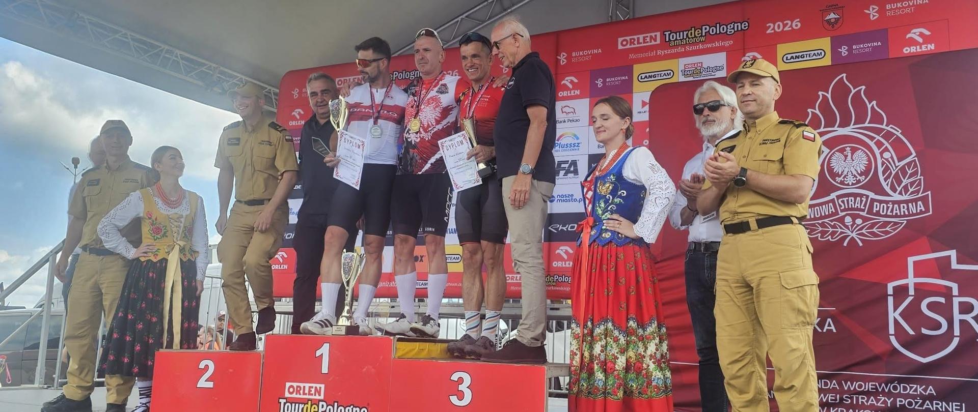 Zdjęcie zrobione po zakończonym wyścigu. Przedstawia zawodników stojących na podium. Zawodnicy stoją w strojach sportowych typowych dla kolarzy. Obok zawodników na podium stoją tez inne osoby – jeden funkcjonariusz Państwowej Straży pożarnej w ubraniu w kolorze piaskowym oraz dwóch mężczyzn. Ponadto zarówno po lewej jak i po prawej stronie obok podium stoją po 3 osoby, w tym po jednej kobiecie w stroju góralskim. Po prawej stronie zdjęcia widoczne jest duże logo Państwowej Straży Pożarnej na czerwonym tle widać czerwoną ściankę, na której są symbole organizatorów oraz sponsorów wydarzenia. Całość znajduje się pod specjalnie przygotowanym zadaszeniem.