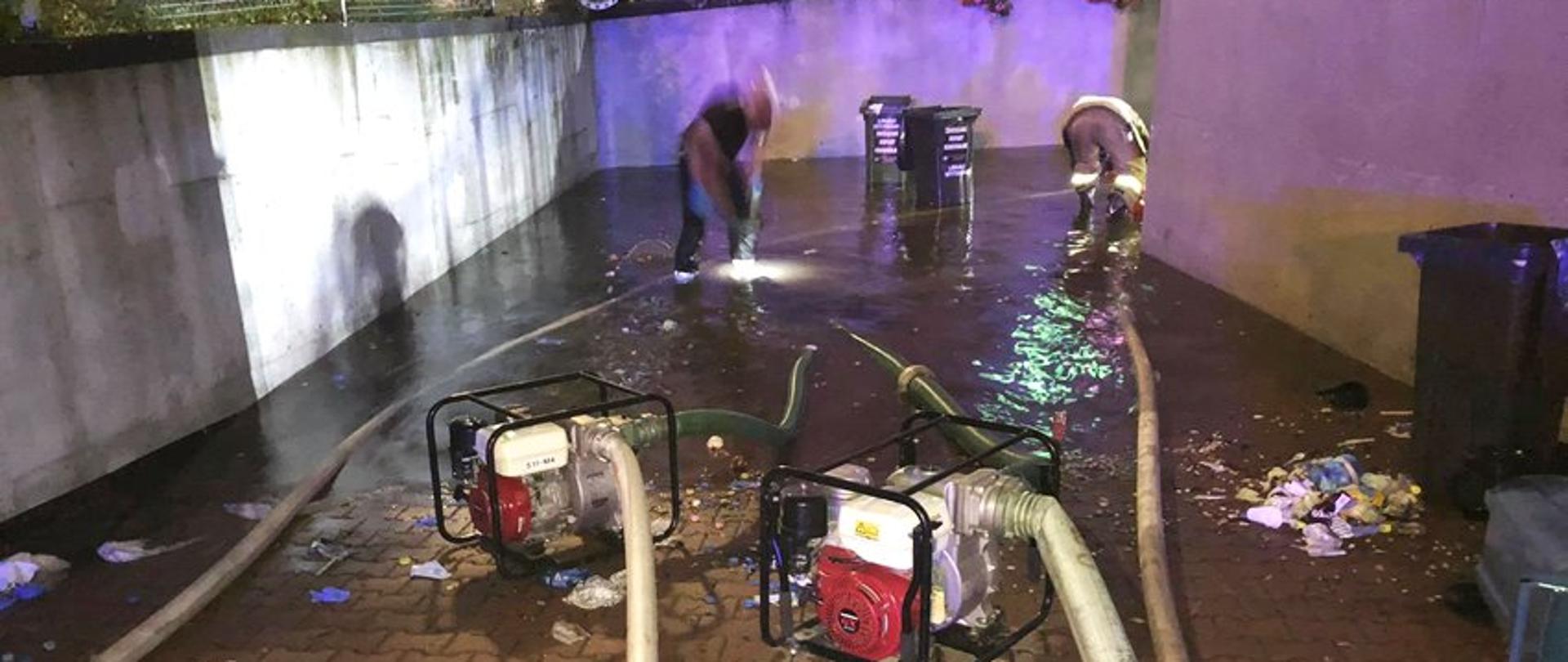 Na zdjęciu widać dwóch strażaków oraz pompę stojących na wjeździe do garażu podziemnego.