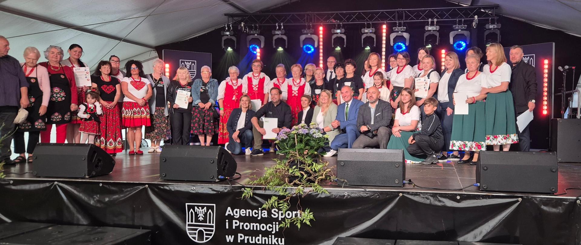 Duża grupa osób pozuje do zdjęcia na scenie pod namiotem. Wśród nich kobiety w strojach regionalnych. Część osób po prawej stronie w pierwszym rzędzie przyklękła do zdjęcia.