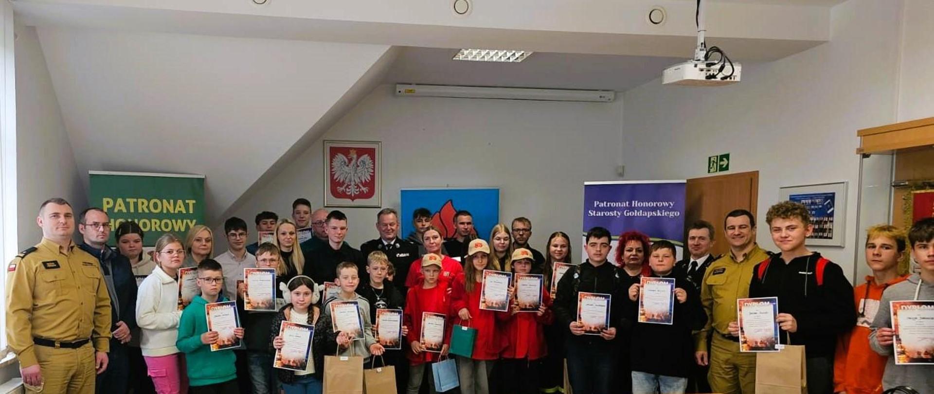 Zdjęcie przedstawia wszystkich uczestników eliminacji powiatowych Ogólnopolskiego Turnieju Wiedzy Pożarniczej „Młodzież Zapobiega Pożarom” z nagrodami/dyplomami oraz opiekunów z poszczególnych szkół i organizatorów przedsięwzięcia, widocznych w świetlicy Komendy Powiatowej Państwowej Straży Pożarnej w Gołdapi