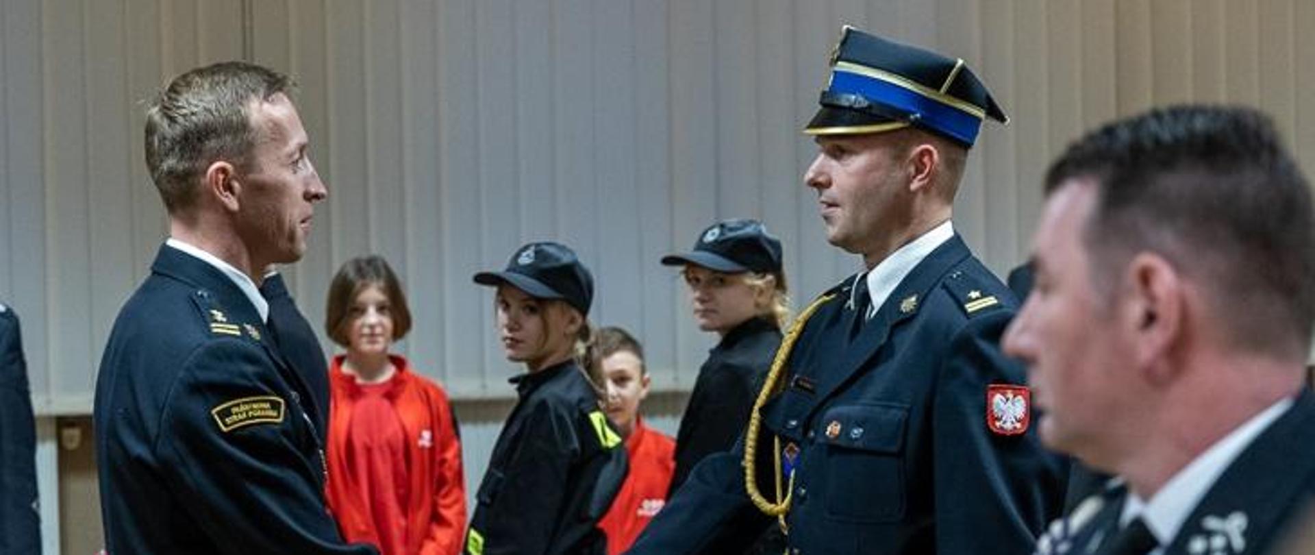 strażak w rogatywce ściska dłoń drugiemu strażakowi, w tle młodzież w mundurach strażackich