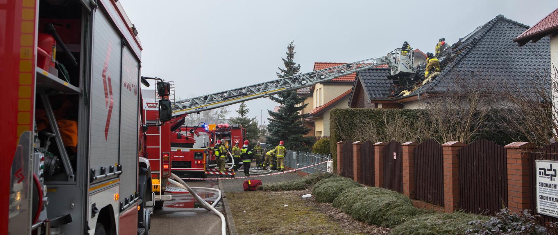 Na zdjęciu widoczni są strażacy oraz wozy bojowe, które prowadzą działania gaśnicze budynku jednorodzinnego. 
