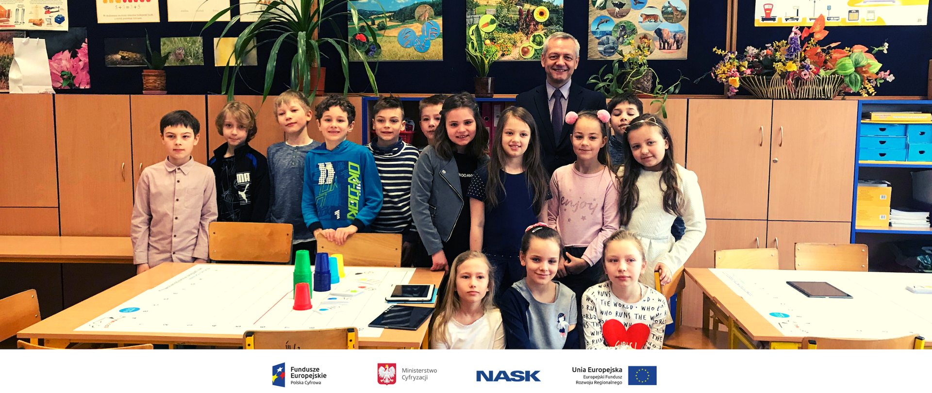 Na zdjęciu widać grupę dzieci z Ministrem Markiem Zagórskim w szkole. Na dole zdjęcia umieszczone są logotypy: Fundusze Europejskie. Polska Cyfrowa, Ministerstwo Cyfryzacji, NASK oraz Unia Europejska. Europejski Fundusz Rozwoju Regionalnego. 