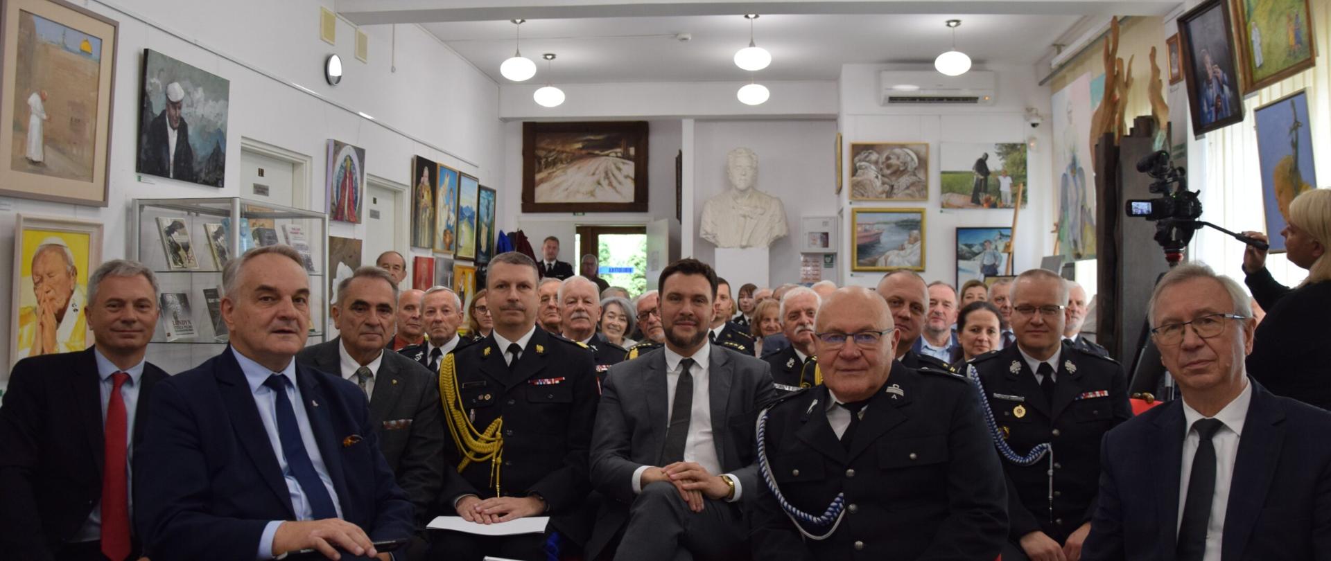 Grupa uczestników wydarzenia siedząca w sali pełnej obrazów i rzeźb. W pierwszym rzędzie widoczni m.in. oficerowie PSP w mundurach galowych oraz osoby w garniturach. W tle liczne portrety i pejzaże, a po prawej stronie operator kamery dokumentujący spotkanie.