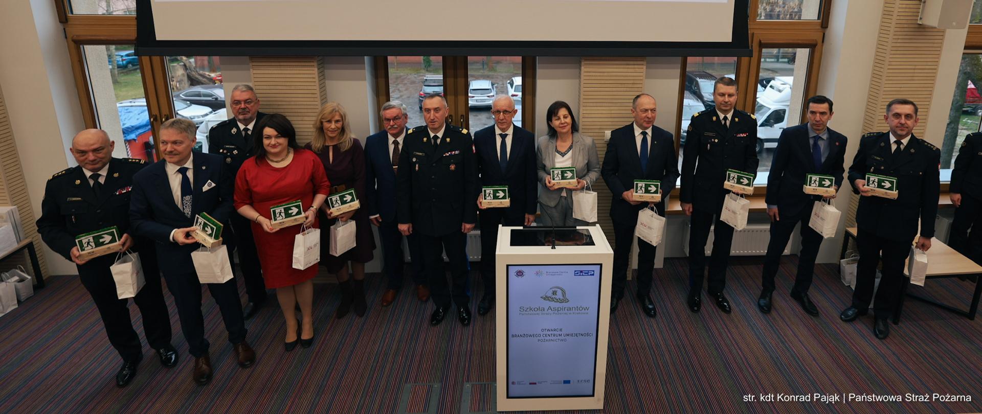 Zdjęcie grupowe gości z pamiątkami.
Ujęcie z góry, z boku, pokazujące grupę gości honorowych i oficerów stojących w rzędzie na scenie. Większość osób (mężczyźni w mundurach i garniturach, kobiety w strojach cywilnych) trzyma w rękach pamiątkowe statuetki z mchem. Pośrodku stoi mównica z logo BCU. Nad nimi znajduje się ekran z napisem „OTWARCIE BRANŻOWEGO CENTRUM UMIEJĘTNOŚCI POŻARNICTWO”.
