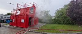 Na zdjęciu widać czerwony kontener w którym aktualnie są przeprowadzane ćwiczenia, przez otwarte drzwi kontenera widać strażaka ubranego w specjalne ubranie trzymającego węża strażackiego, który jest napełniony wodą. Z kontenera wydobywa się dym.