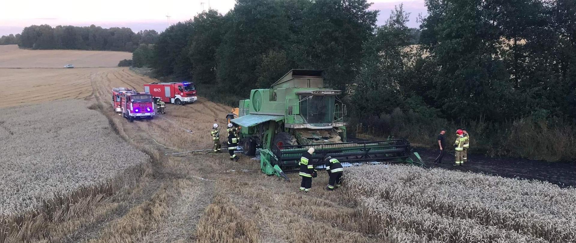 Na ściernisku stoi kombajn, strażacy którzy dokonywali jego gaszenia oraz wozy bojowe