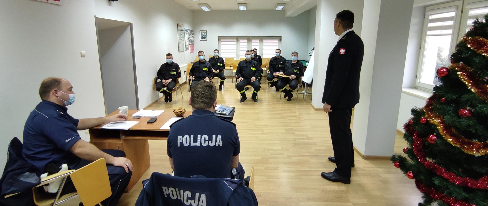 na pierwszym planie siedzący tyłem Policjanci przy biurku, po prawej Choinka a przed nią tyłem do obiektywu Komendant Powiatowy w umundurowaniu galowym. w tle strażacy w umundurowaniu koszarowym w maseczkach. Spotkanie szkoleniowe w świetlicy KP PSP w Ropczycach