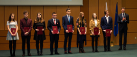 Najzdolniejsi uczniowie stoją z ministrem edukacji na scenie. W tle fragment prezentacji, flaga Polski i Unii Europejskiej.